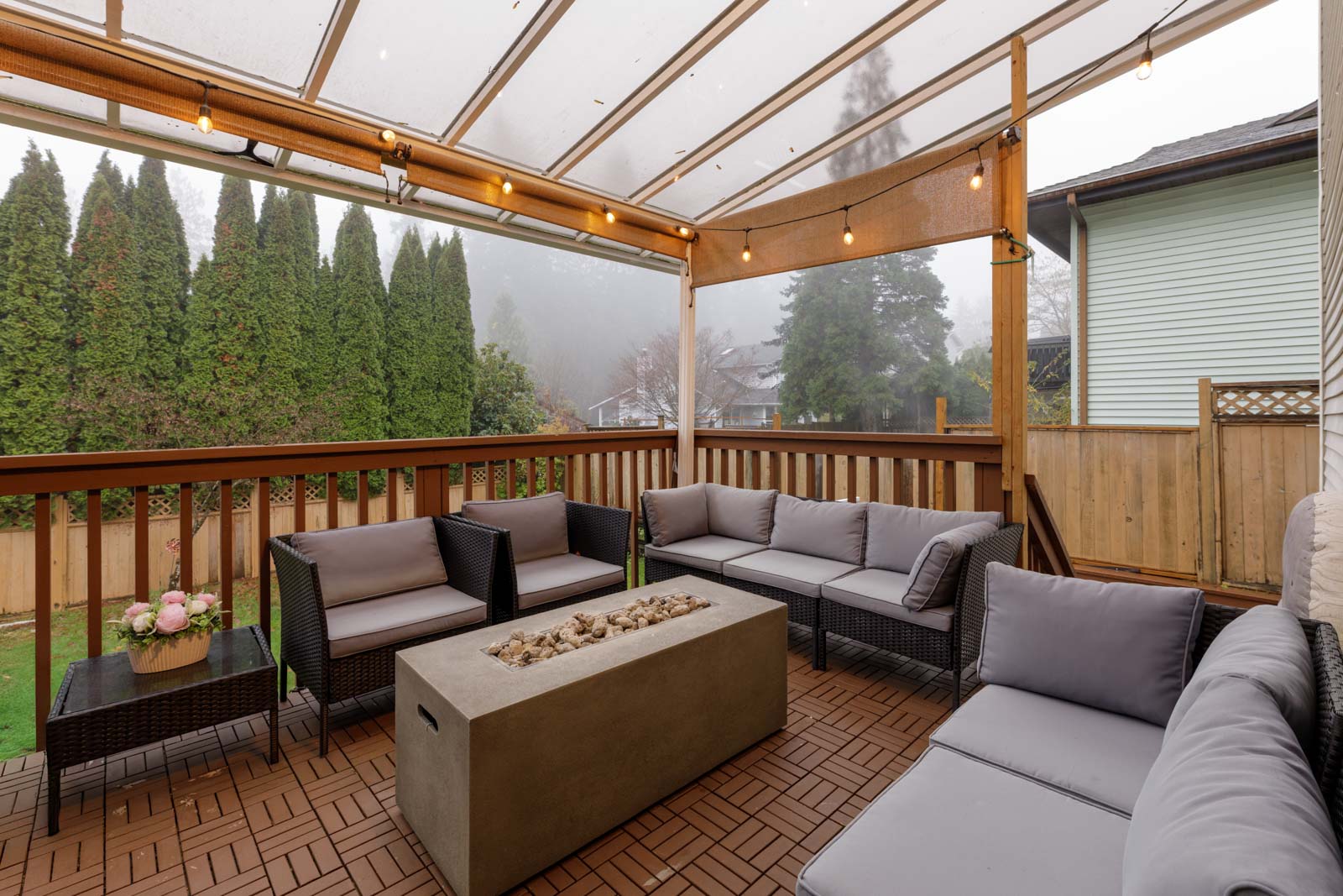 Covered patio with string lights, gray cushioned outdoor seating, concrete fire pit, wooden deck, and view of trees and neighboring houses in the background.