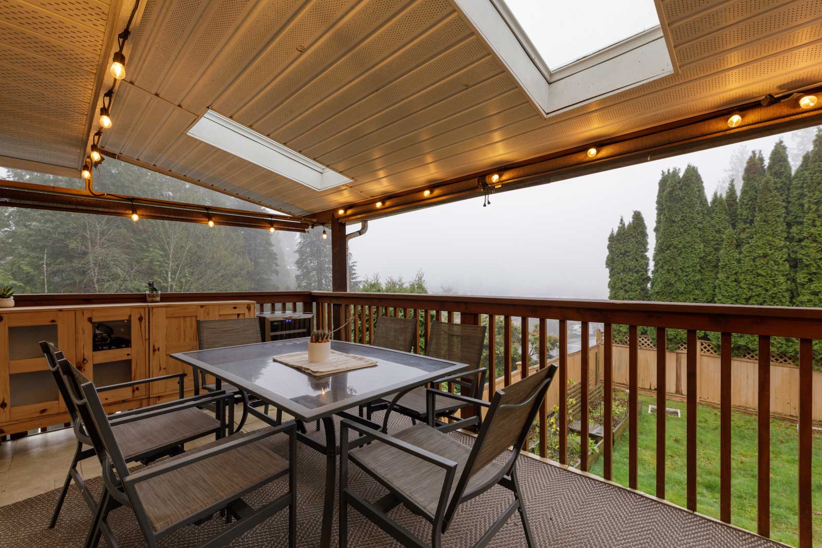 Covered patio with string lights, a skylight, and an outdoor dining table with four chairs, overlooking a fenced backyard and tall evergreen trees on a foggy day.