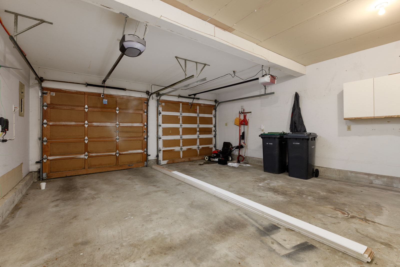 A clean, empty two-car garage with wooden doors, two trash bins, a lawnmower, a fire extinguisher on the wall, and a white cabinet; long boards lie on the concrete floor.