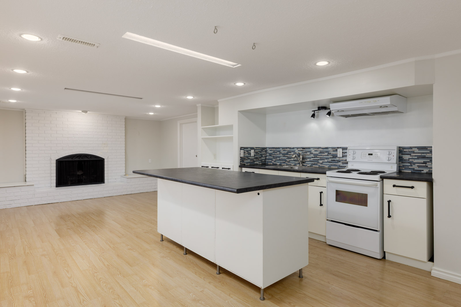 Modern kitchen with white cabinets, a black countertop island, white appliances, tile backsplash, and light wood flooring, adjacent to a living area with a white brick fireplace.