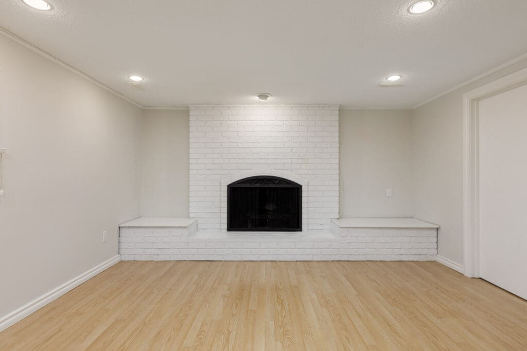 A white brick fireplace with a black grate is centered on a beige wall, flanked by two built-in benches. The room has recessed lighting and light wood flooring.