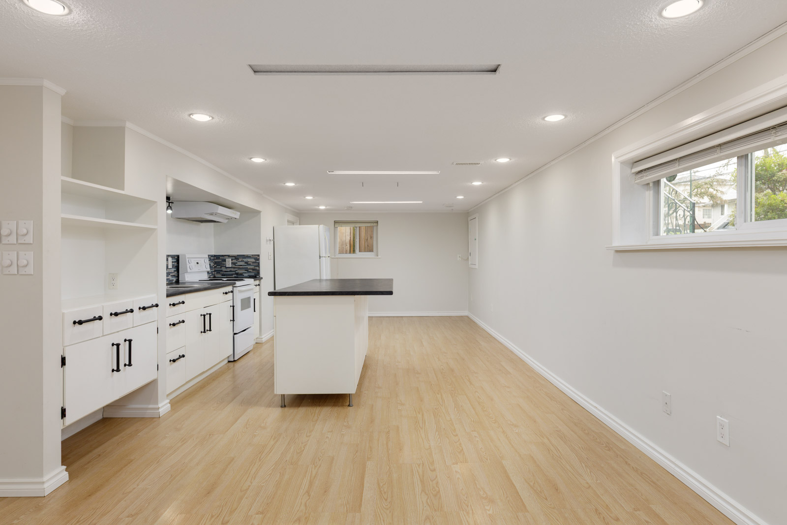 Bright basement kitchen with white cabinets, center island, wood flooring, and a window letting in natural light. The space is empty and features recessed lighting.