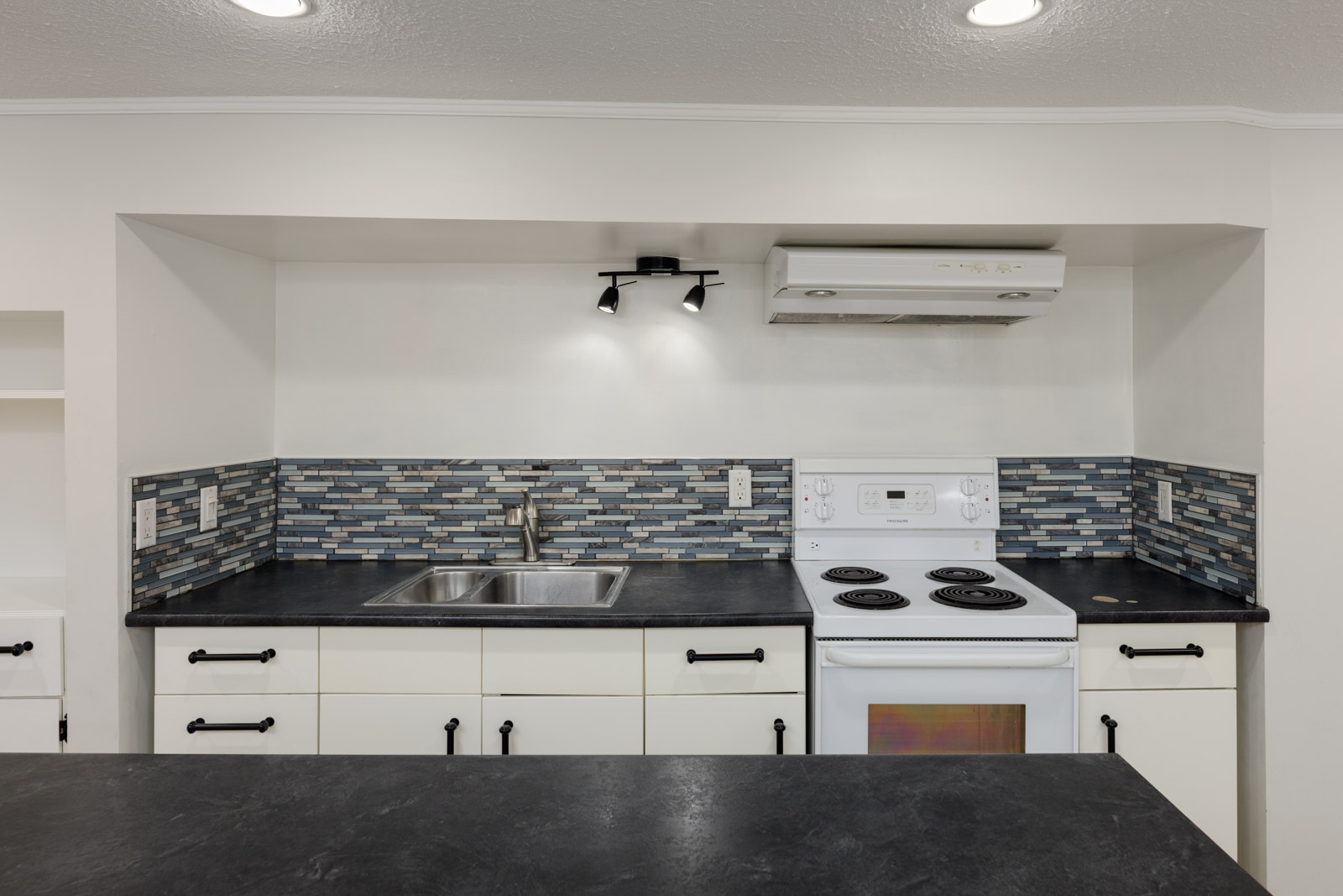 A modern kitchen with white cabinets, black countertops, a stainless steel sink, an electric stove, range hood, and blue-gray mosaic tile backsplash.