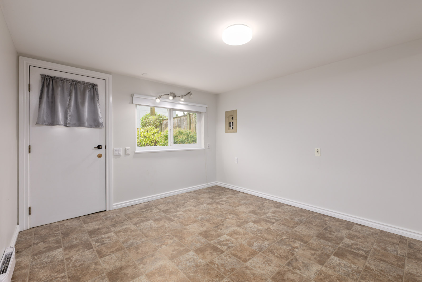 Empty room with beige vinyl tile flooring, white walls, a window with a view outside, a door with a gray curtain, and an overhead ceiling light.