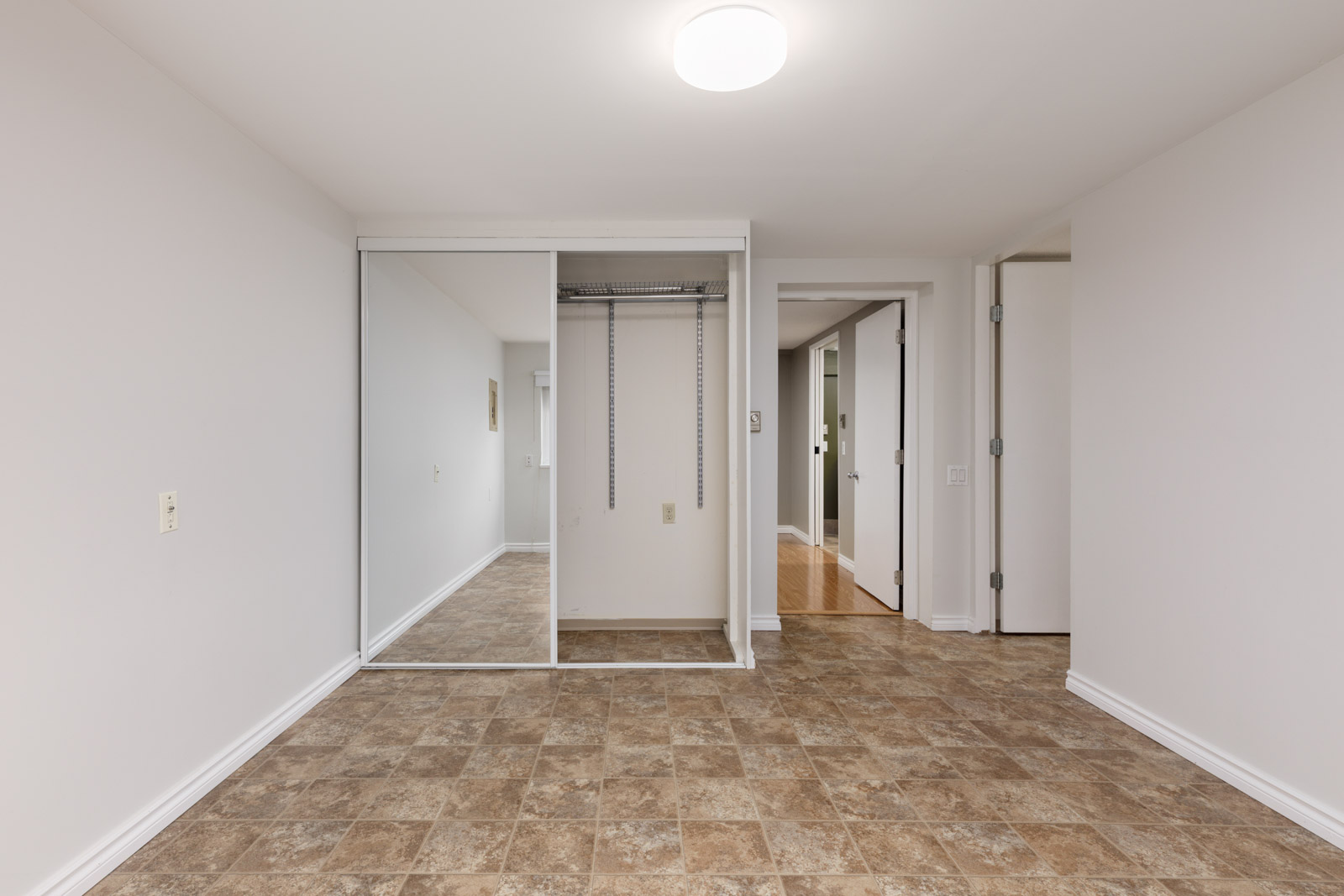 Empty room with tiled floor, white walls, a ceiling light, and a closet with mirrored sliding doors. The room has open doorways leading to a hallway.