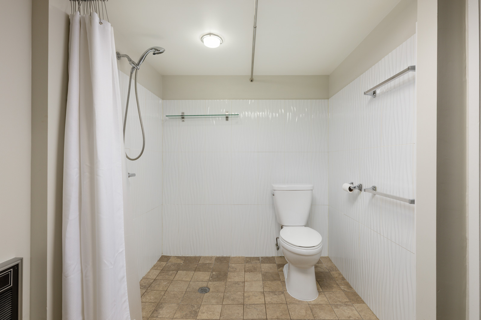 Accessible bathroom with walk-in shower, white curtain, wall-mounted grab bars, toilet, beige tile floor, and overhead light.