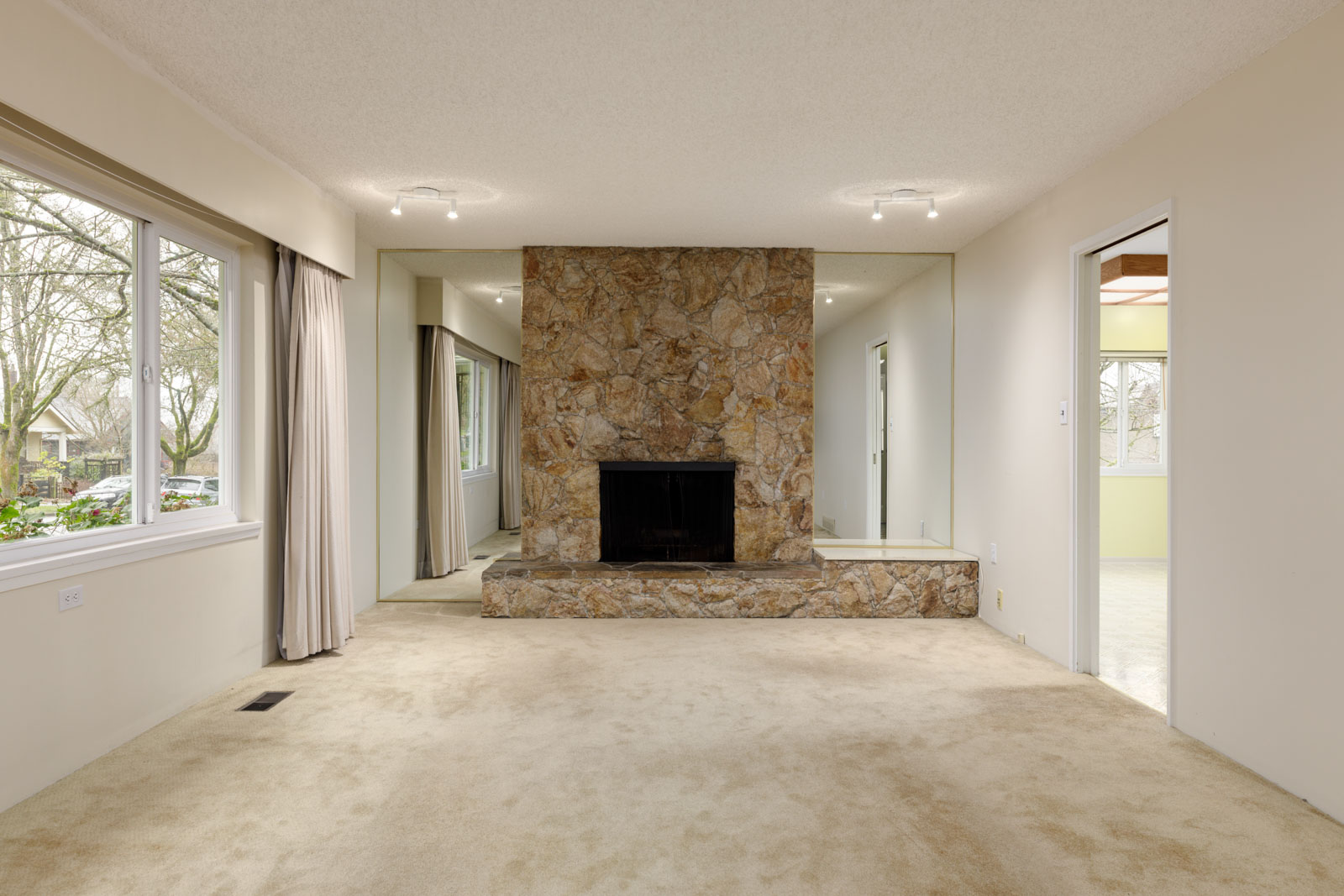 A living room with beige carpet, a stone fireplace, mirrored walls, and large windows with a view of trees.