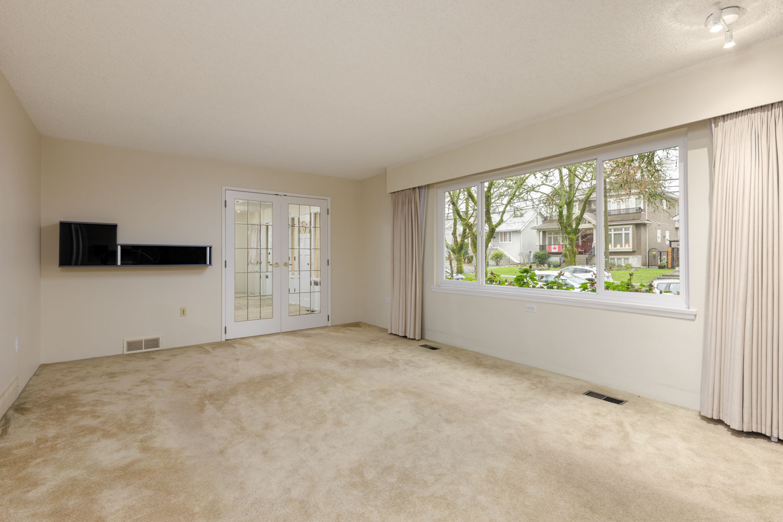Empty living room with beige carpet, large window with curtains, wall-mounted shelves, and glass double doors leading to another room.