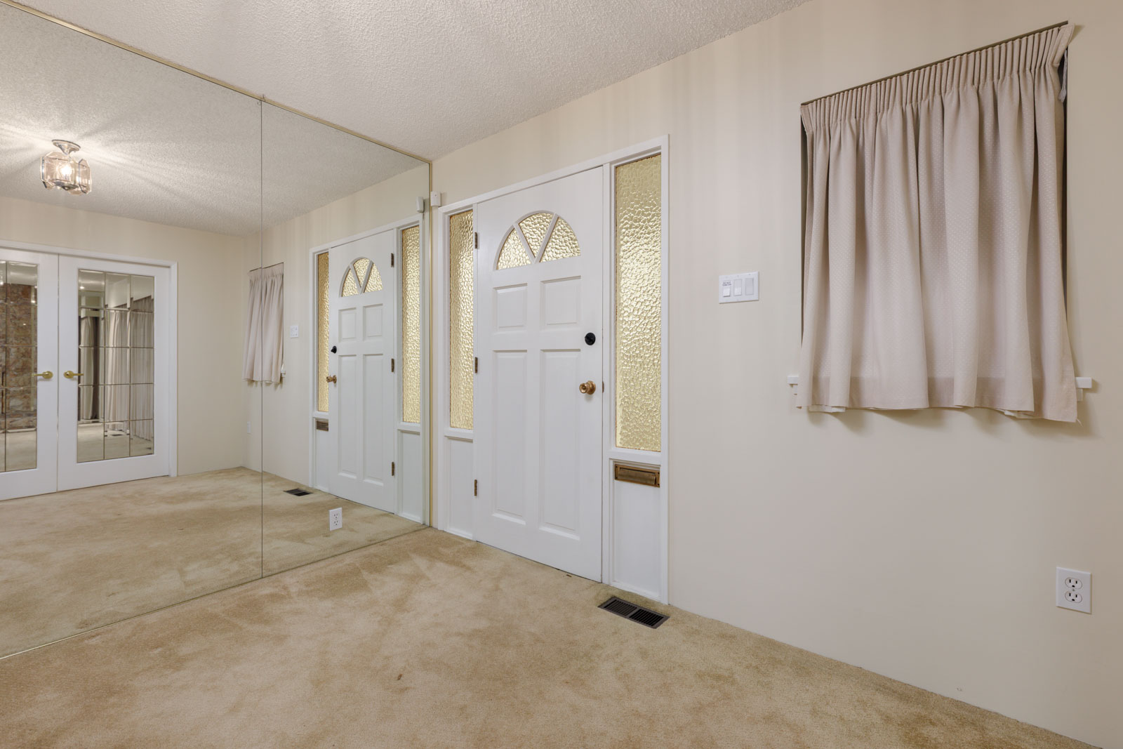 A beige-carpeted entryway with a white front door, decorative glass panels, mirrored wall, and a window with beige curtains.