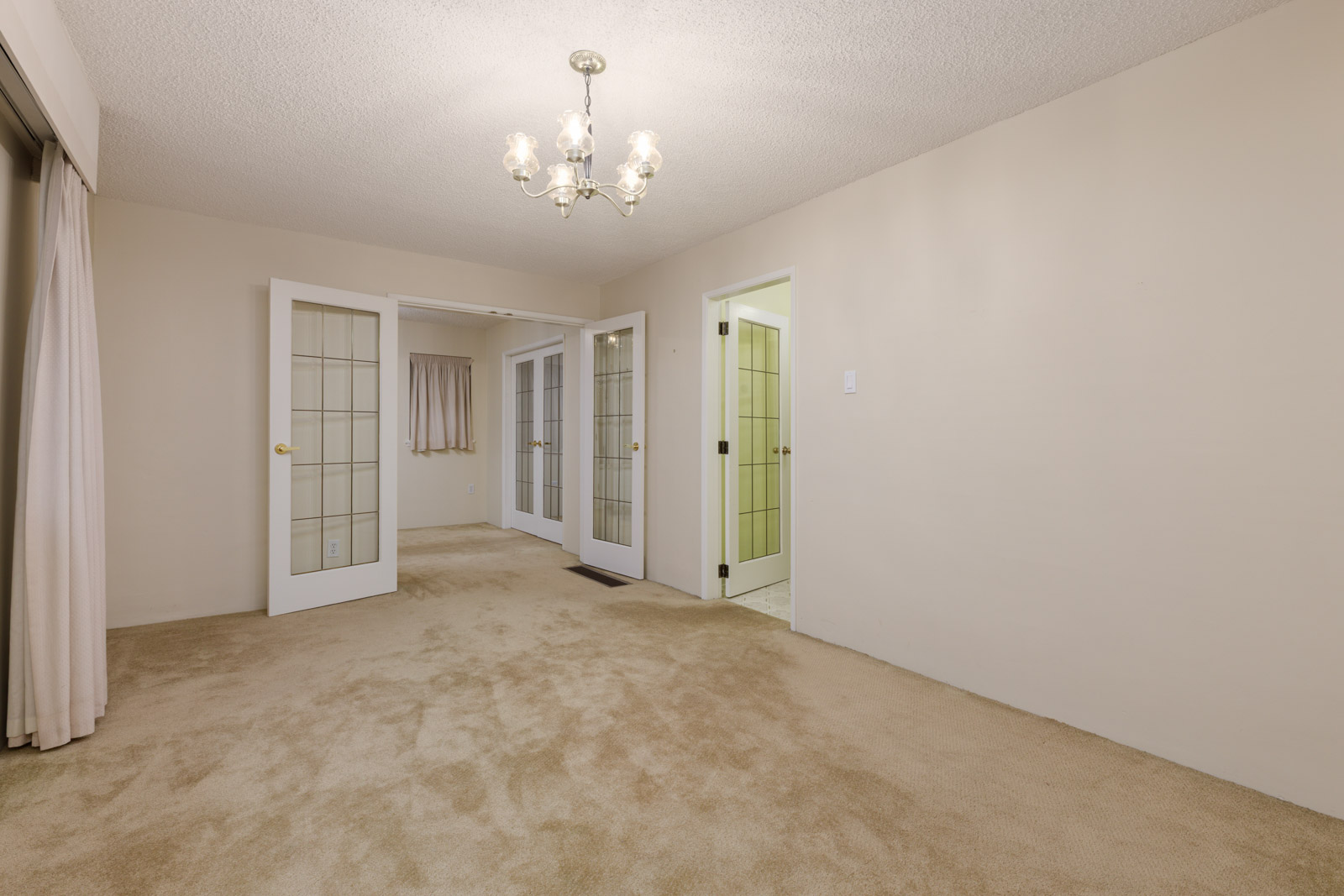 Empty room with beige carpet, cream walls, a chandelier, two sets of glass French doors, a doorway to a tiled room, and a window with beige curtains.