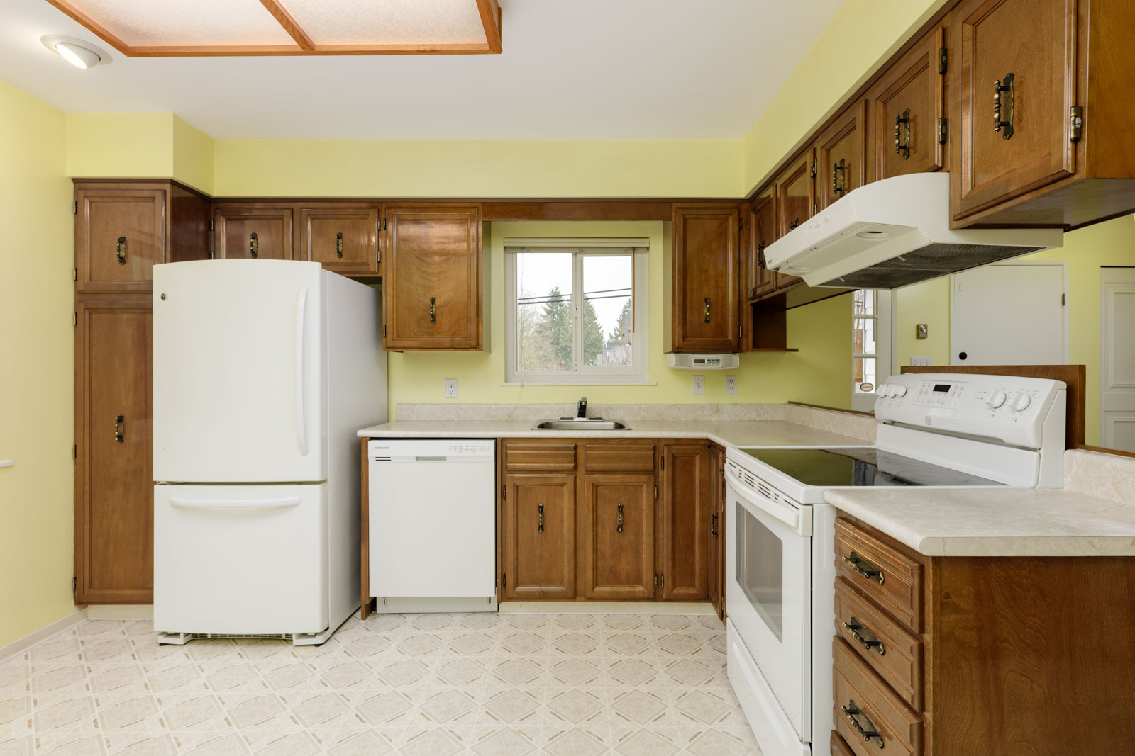 A kitchen with wooden cabinets, white appliances including a refrigerator, dishwasher, and stove, a window above the sink, and yellow walls.