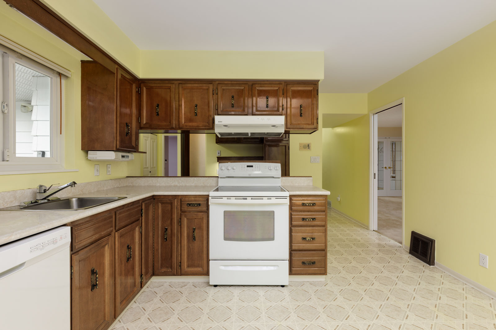 Kitchen with brown wooden cabinets, white electric stove, beige countertops, dishwasher, and yellow walls; patterned linoleum flooring and a window above the sink.