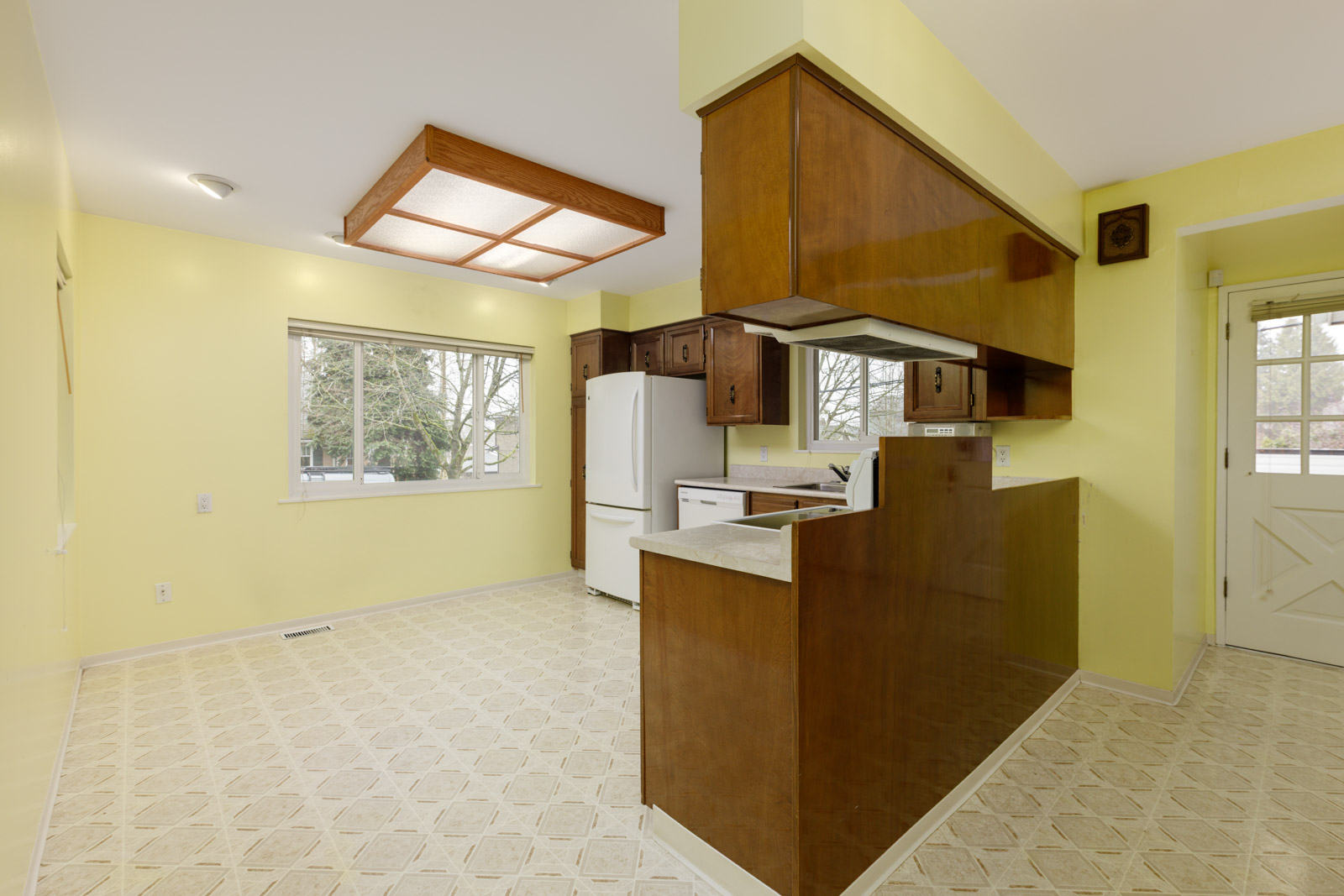 Kitchen with yellow walls, wooden cabinets, white appliances, a patterned linoleum floor, and large window overlooking trees. A door is visible on the right side.