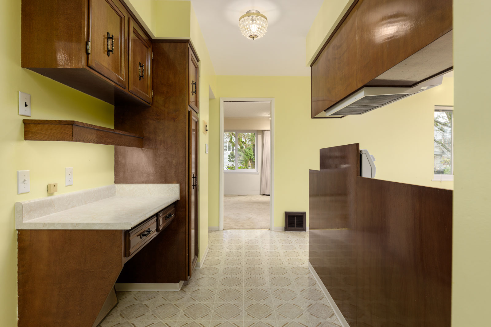Narrow kitchen with yellow walls, wood cabinets, white countertops, patterned linoleum floor, and a view into a carpeted living room with a window.