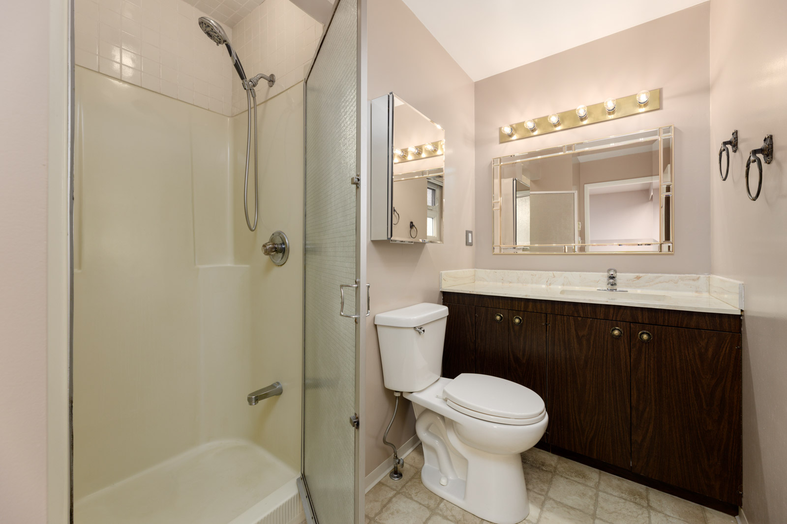 Bathroom with a glass shower, white toilet, brown vanity with marble countertop, large mirror, and overhead lights.