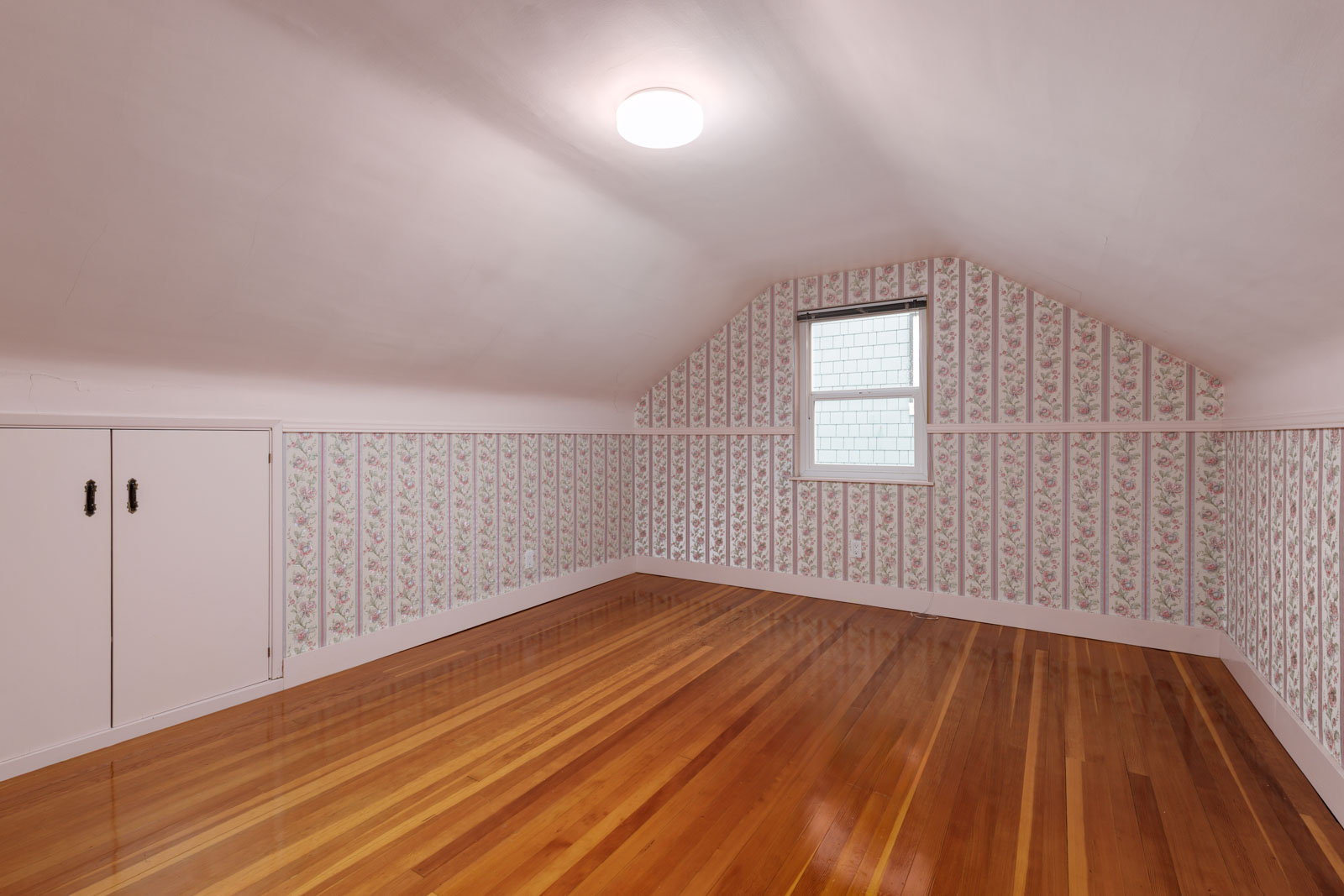 Empty attic room with floral wallpaper, a single window, overhead light, glossy wooden floor, and small built-in storage doors on the left wall.