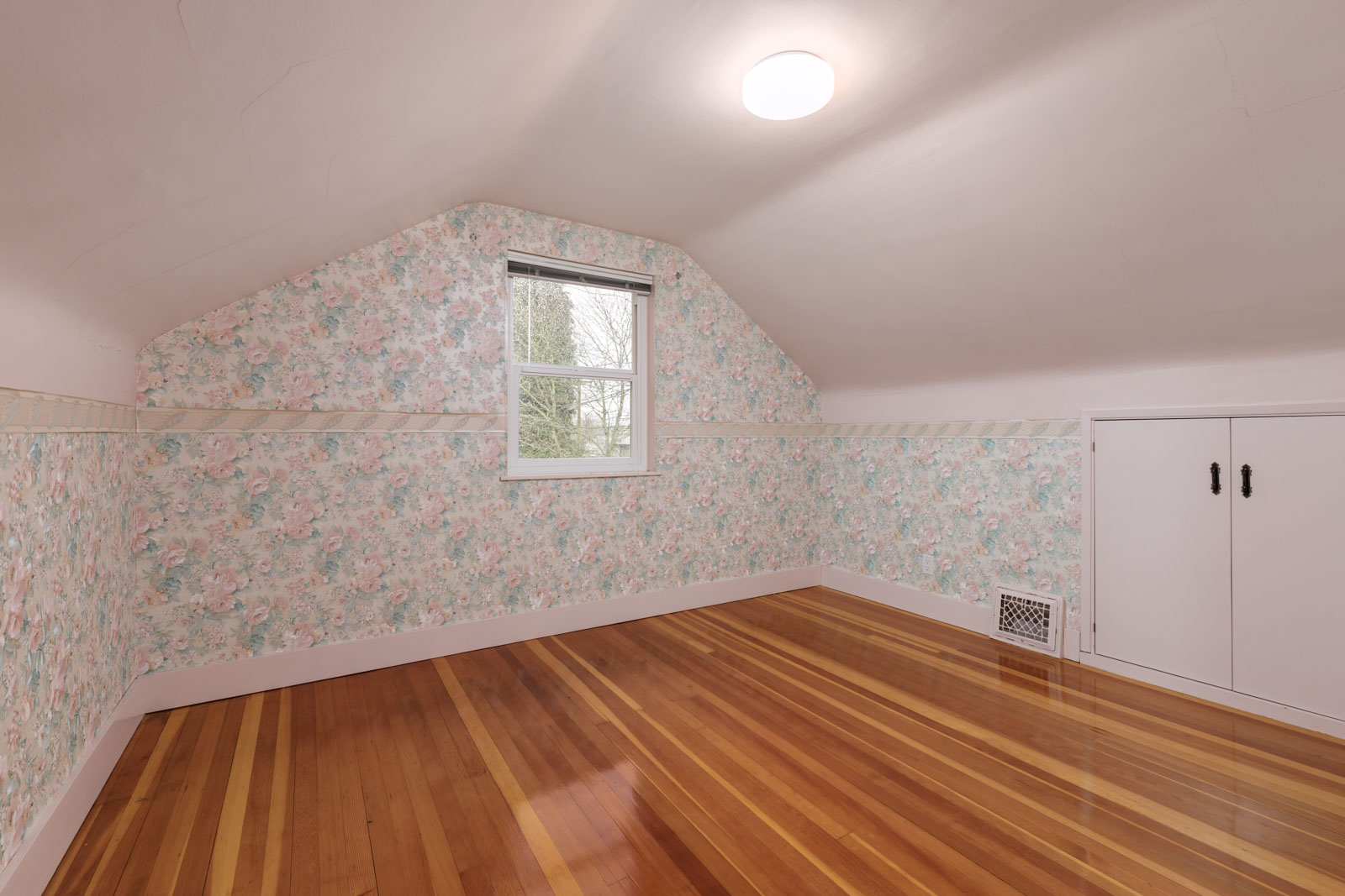 Empty attic room with floral wallpaper, a single window, wood flooring, sloped ceiling, a ceiling light, and a small built-in cabinet on the right wall.