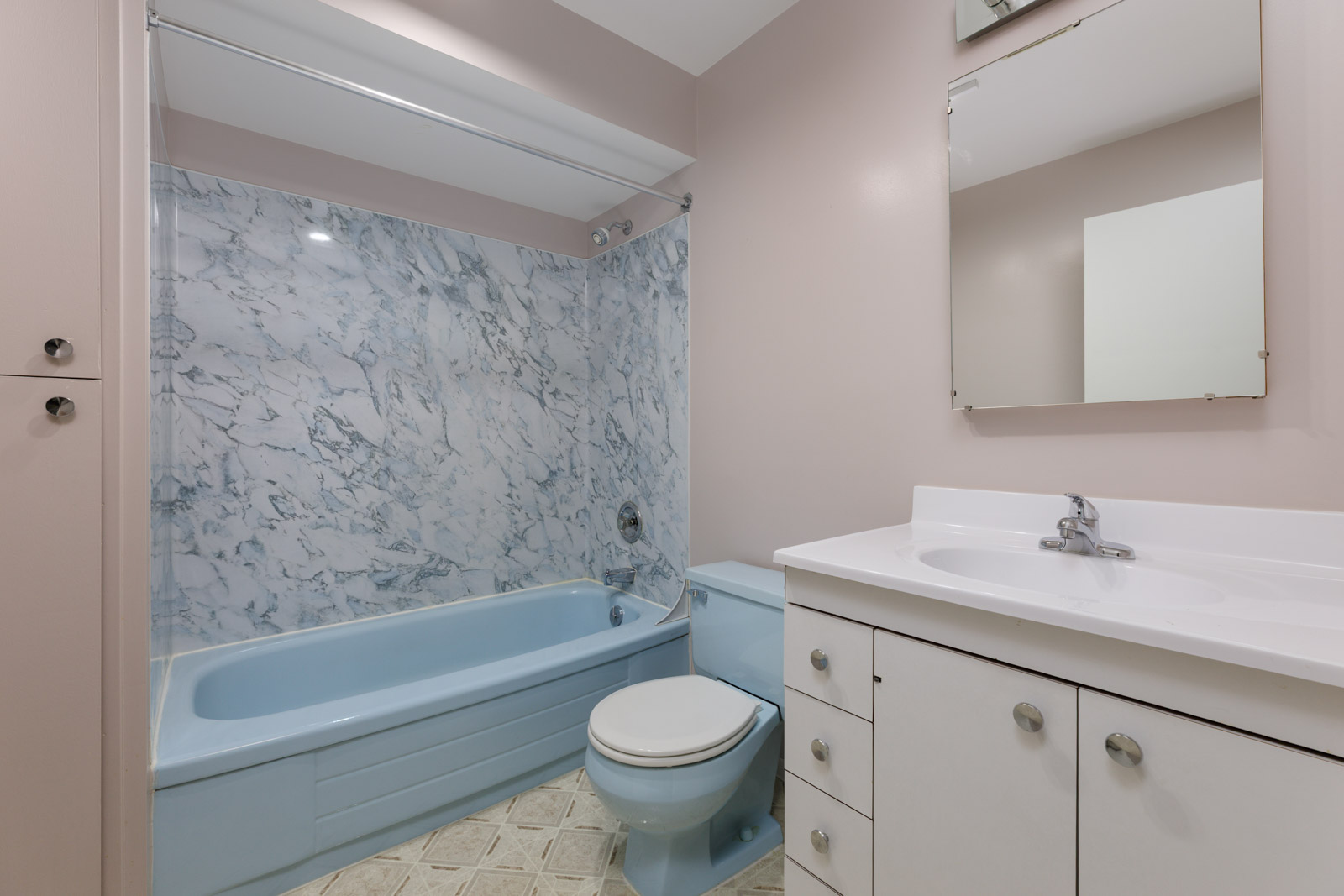 Bathroom with light pink walls, a blue bathtub and toilet, marble-patterned shower wall, white vanity with sink, and a mirror above the sink.