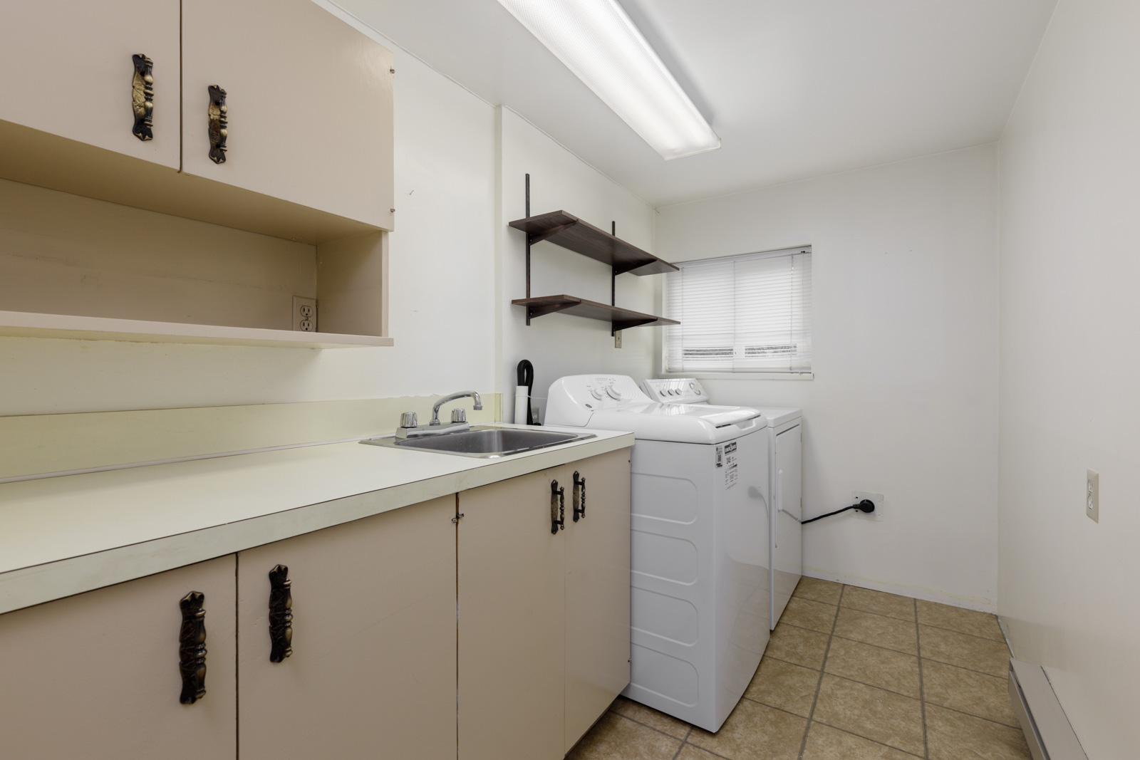 Laundry room with beige cabinets, a sink, white washing machine and dryer, wall shelves, tiled floor, and a window with blinds.