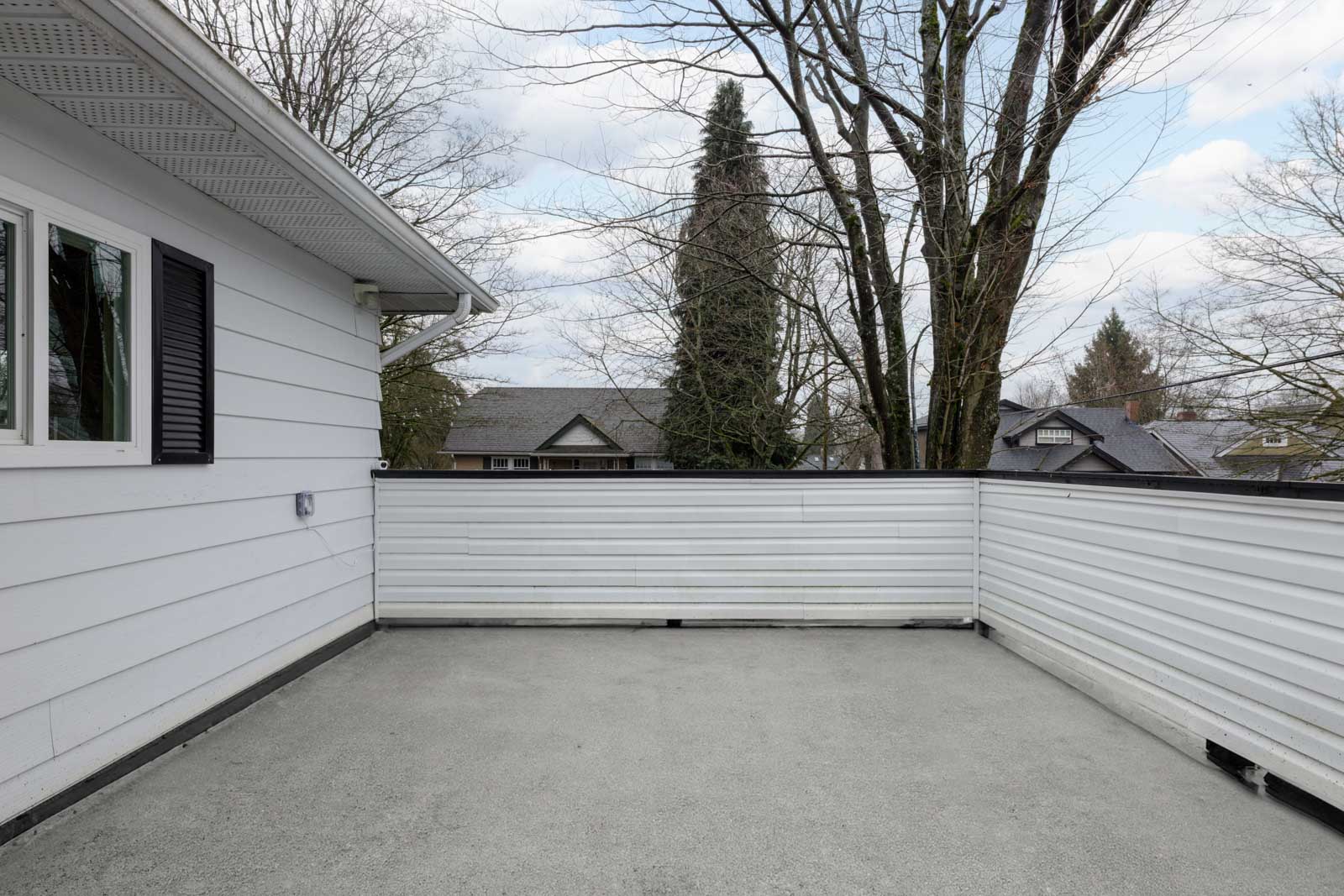 A rooftop deck with a light gray floor and white railing, attached to a white house. Leafless trees and neighboring houses are visible in the background under a cloudy sky.
