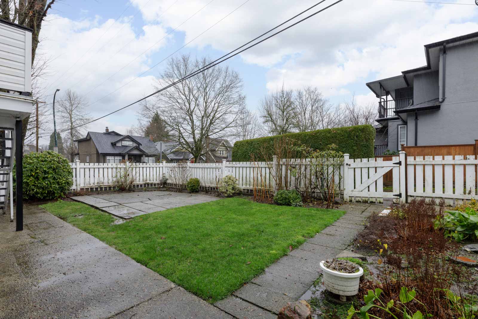 A small backyard with a green lawn, paved walkway, white picket fence, and some potted plants on a cloudy day.