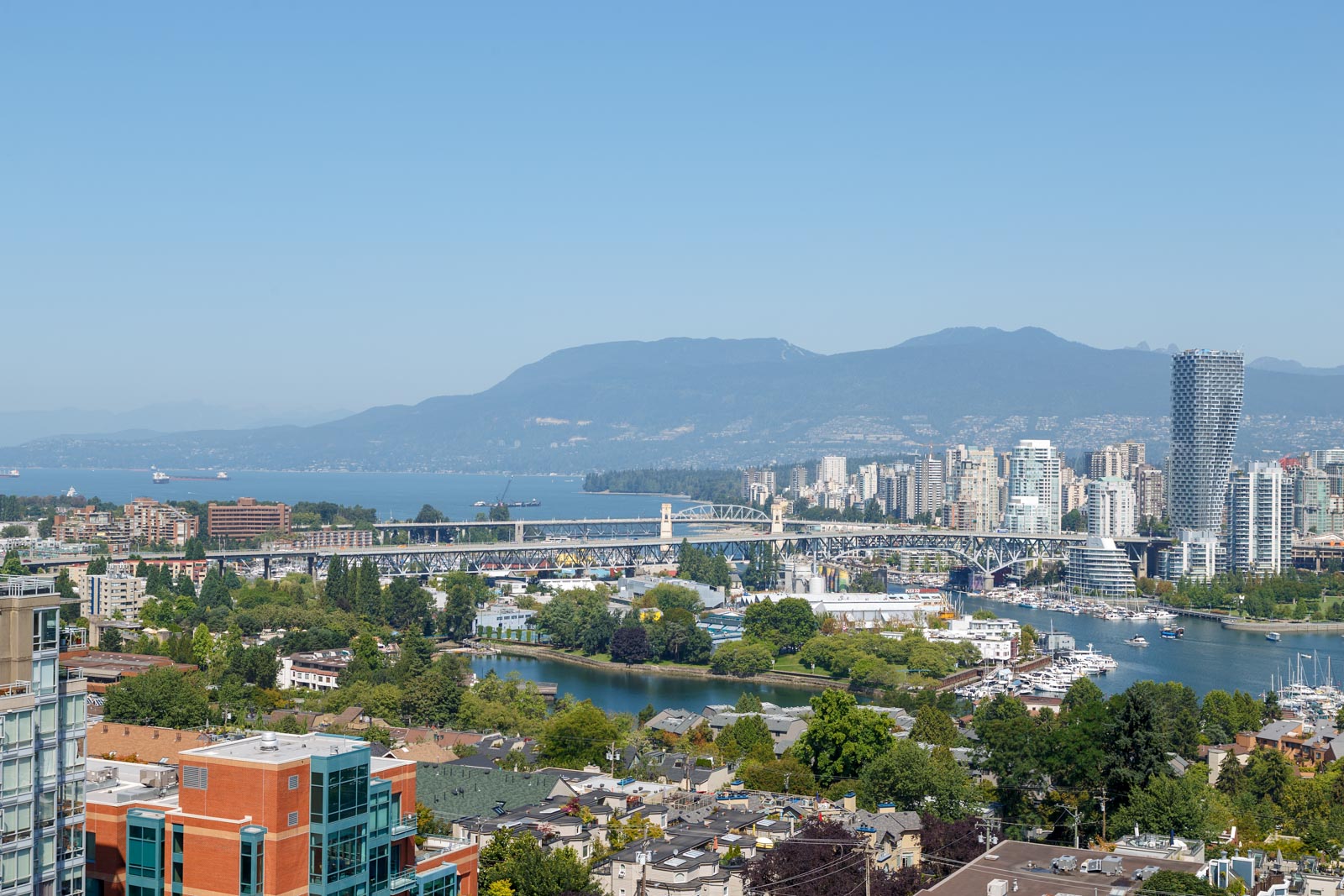 A cityscape featuring a bridge over a body of water, with buildings, greenery, and mountains in the background under a clear blue sky.