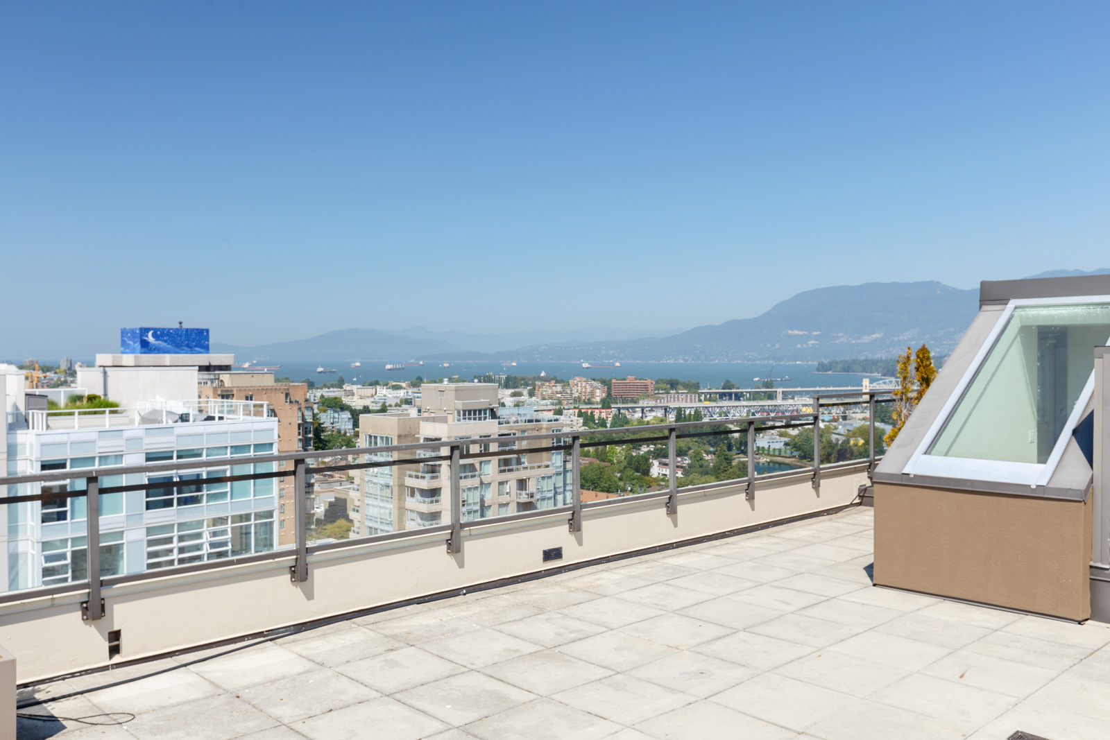 Rooftop terrace with glass railings overlooking city buildings, water, and distant mountains under a clear blue sky.