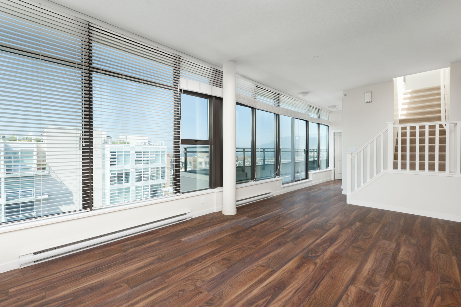 Empty modern living room with large windows, wooden flooring, white walls, and a staircase leading to an upper floor.