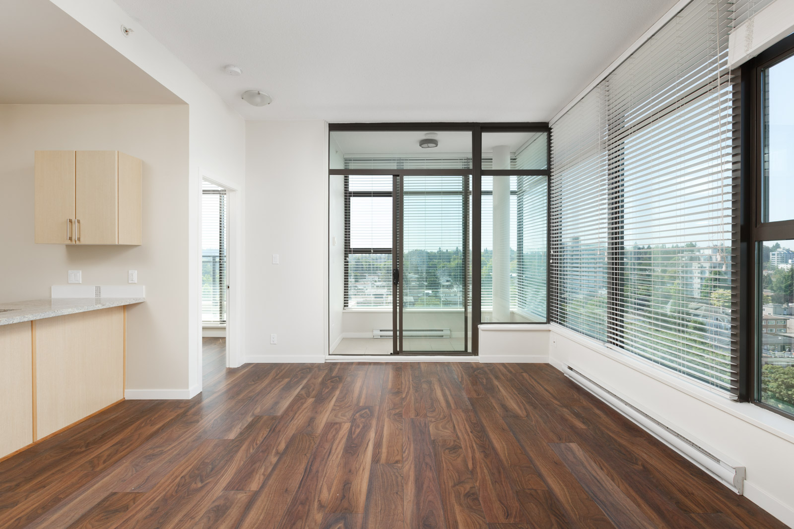 Modern empty apartment with large windows, wood flooring, white walls, and a small balcony enclosed with glass panels overlooking a city view.