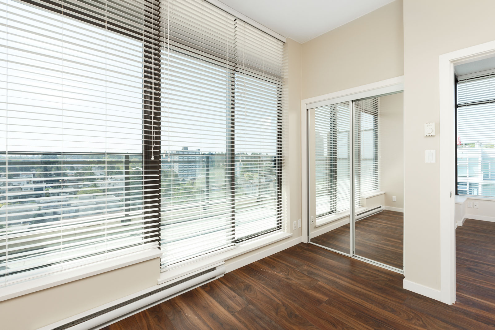 A bright, empty room with large windows, white blinds, mirrored closet doors, and dark wood flooring, overlooking a cityscape.