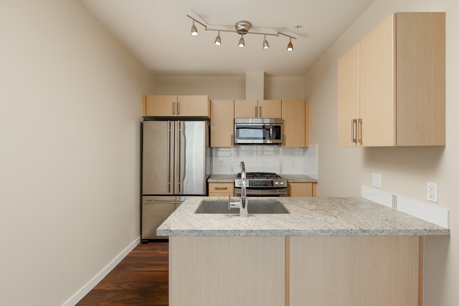 Modern kitchen with light wood cabinets, stainless steel appliances, granite countertop, and under-mount sink; track lighting on ceiling.