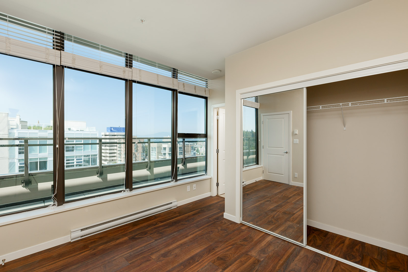 Empty room with large windows, wood flooring, a mirrored closet, and a view of city buildings and blue sky outside.