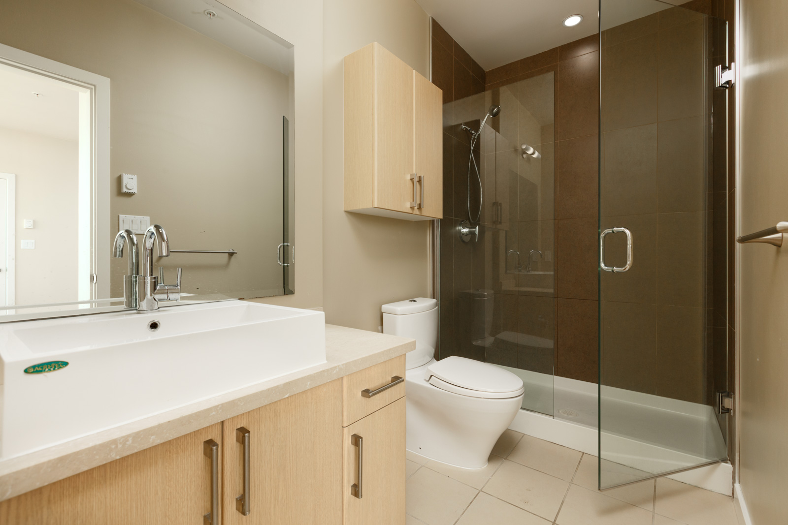 Modern bathroom with a rectangular sink, light wood cabinets, a toilet, and a glass-enclosed shower with brown tiles.