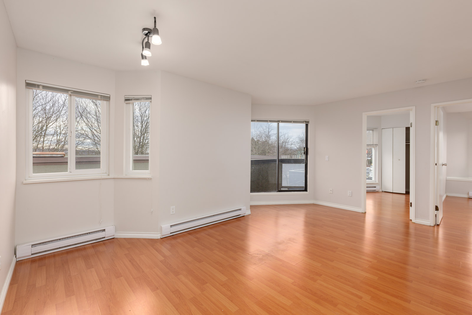 Unfurnished apartment room with light walls, wood flooring, large windows, baseboard heaters, and two doorways leading to adjacent rooms.