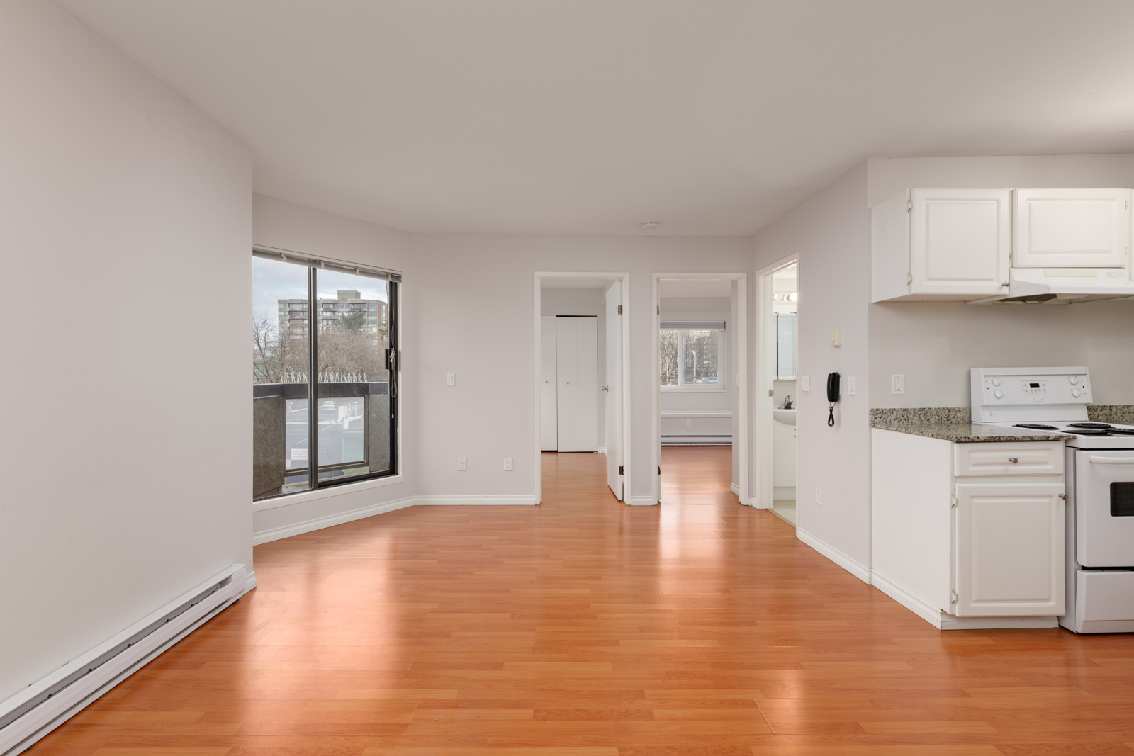 Unfurnished apartment with white walls, hardwood floors, an open kitchen, large windows, and a balcony view. Multiple doorways lead to other rooms.