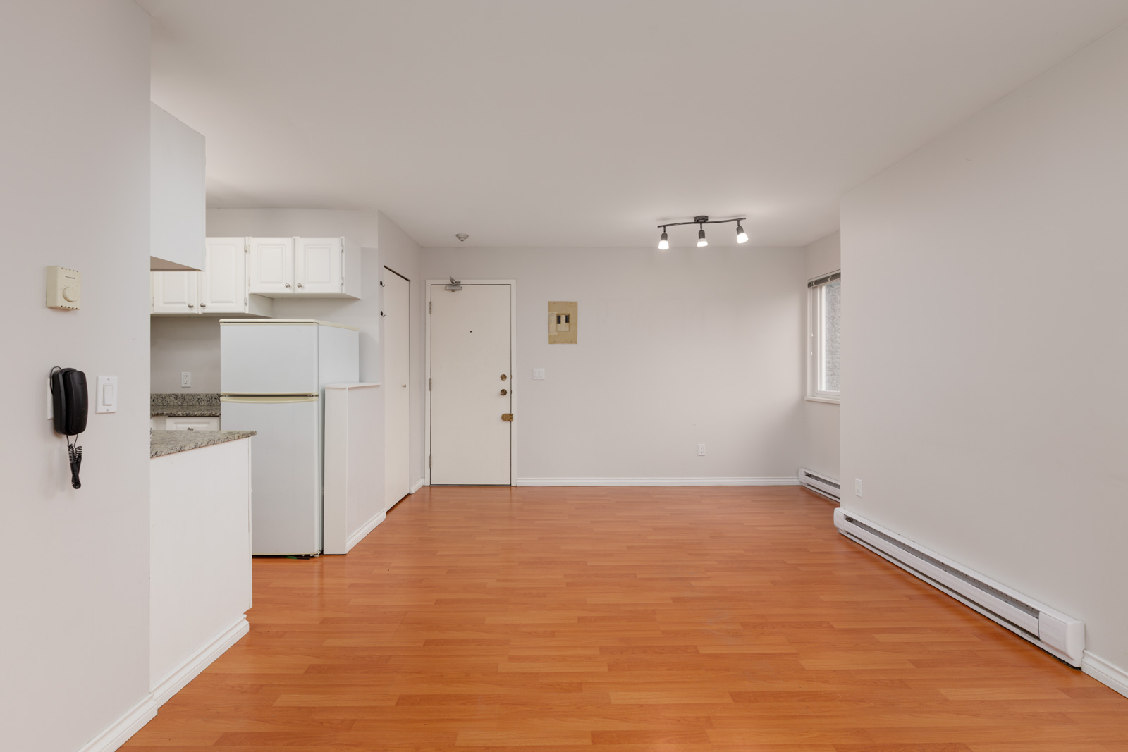 Unfurnished apartment with wood flooring, white walls, a small kitchen area, white cabinets, a refrigerator, entrance door, and a window with blinds.