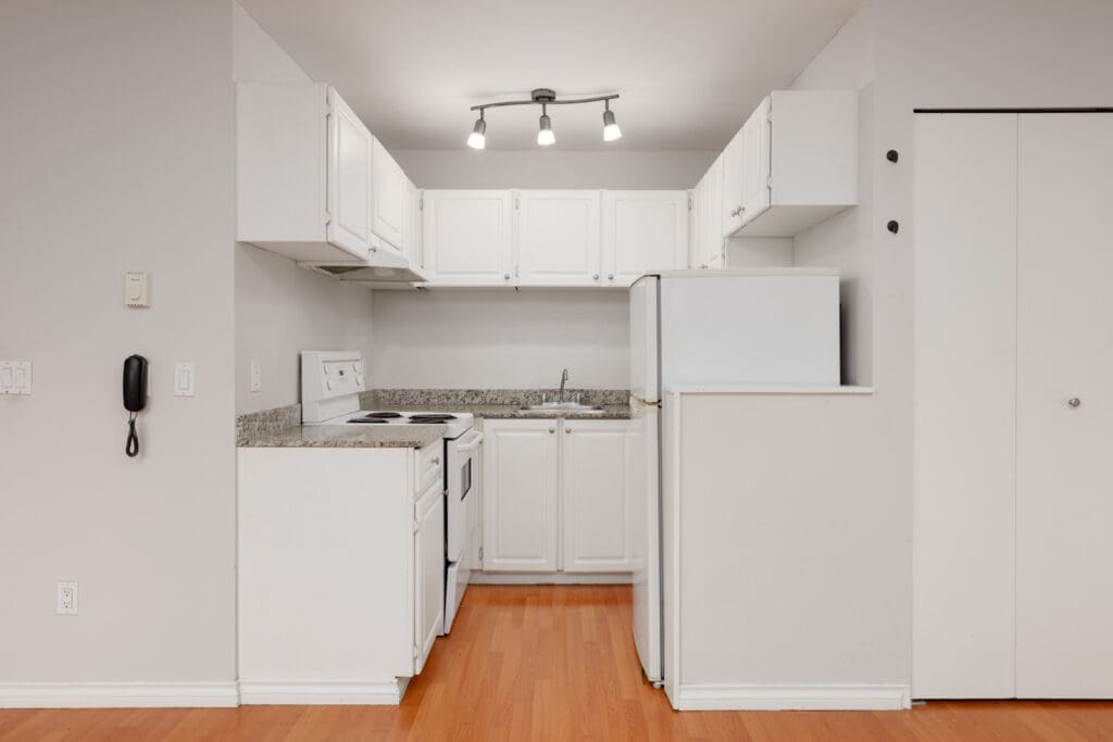 Small kitchen with white cabinets, white appliances, granite countertop, hardwood floor, and track lighting. A closed closet door is on the right side.