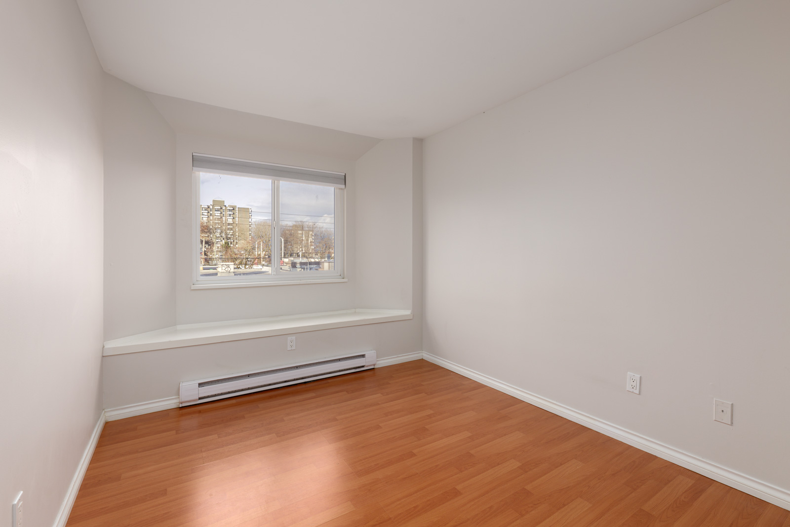 Empty room with light-colored walls, a large window with a view of buildings, wood flooring, a white baseboard heater, and a built-in ledge under the window.