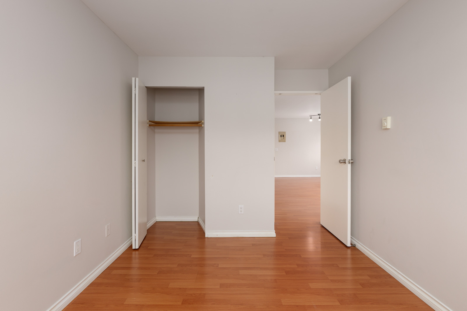 Empty room with light gray walls, wood laminate flooring, an open closet with a shelf and rod, and an open door leading to another room.