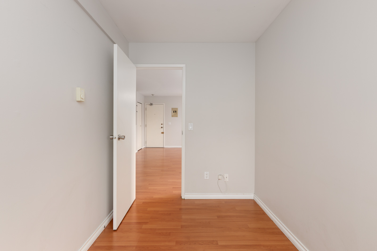 A small, empty room with light gray walls, an open white door, and hardwood floors, leading to a larger room with similar flooring.