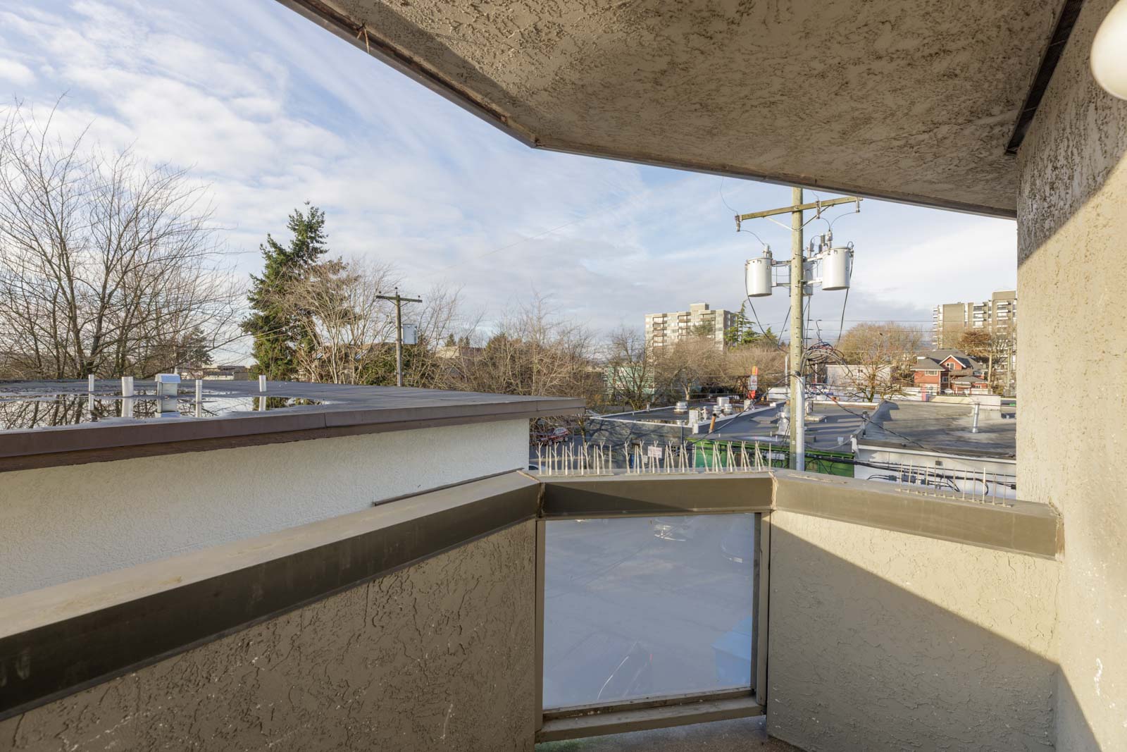 View from a small balcony with concrete walls, overlooking trees, power lines, rooftops, and distant apartment buildings under a partly cloudy sky.