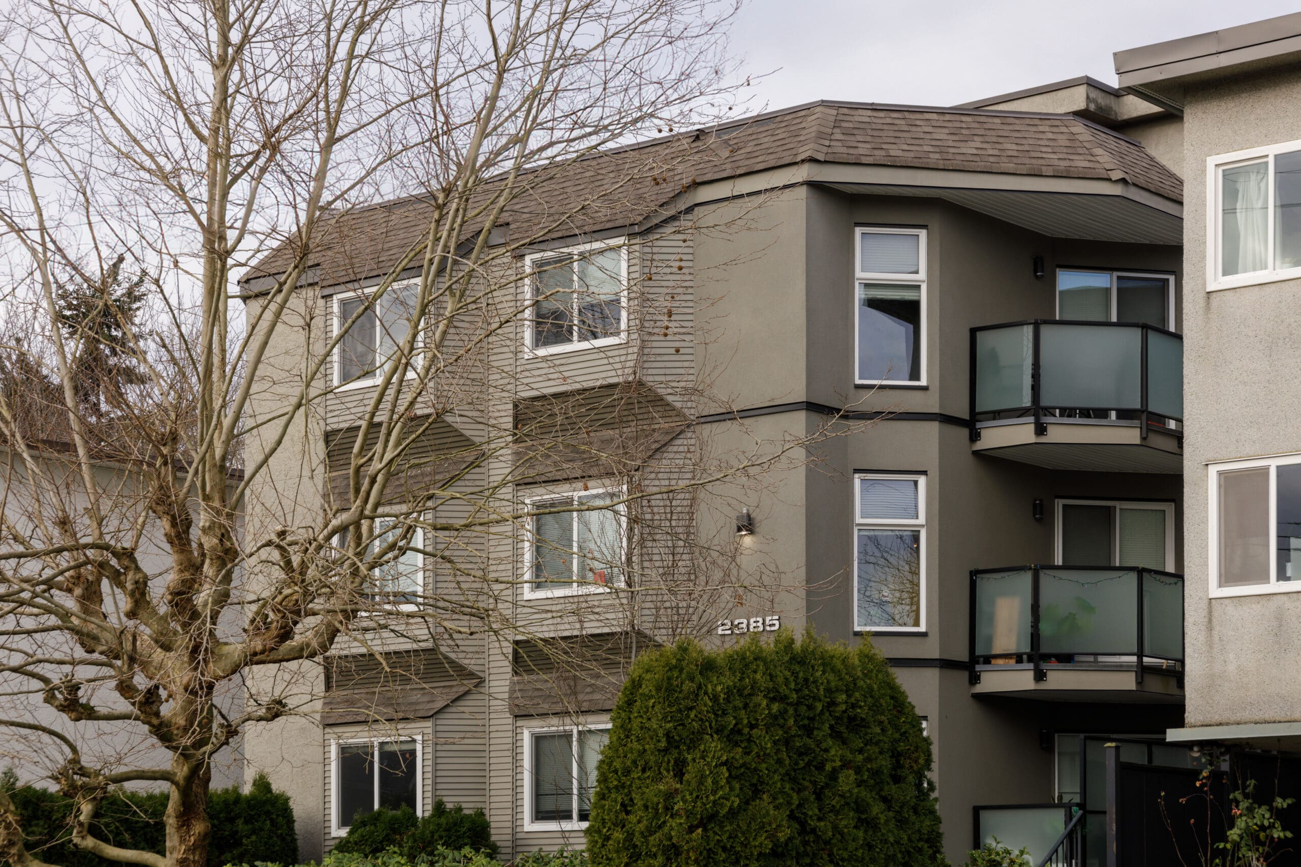 A three-story apartment building with beige and gray exterior, balconies with glass railings, and a leafless tree in front.