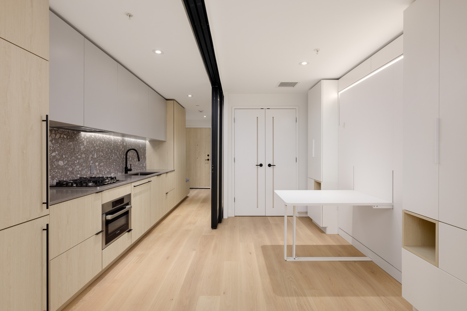 Modern minimalist kitchen with light wood cabinets, gray backsplash, built-in appliances, and a fold-down white table in a clean, bright apartment interior.