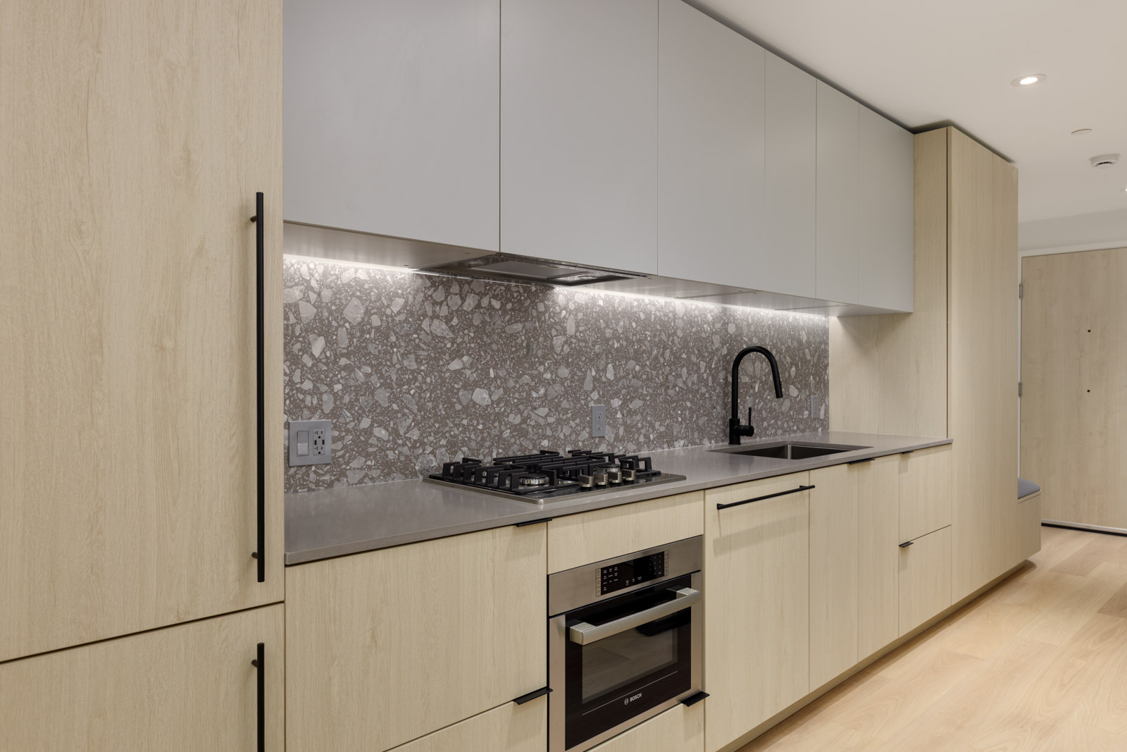 Modern kitchen with light wood cabinets, built-in oven, gas cooktop, gray terrazzo backsplash, under-cabinet lighting, and a black faucet over a gray countertop.