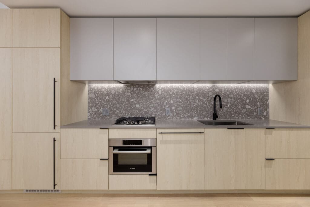 Modern kitchen with light wood cabinets, gray countertops, built-in oven, gas stove, gray upper cabinets, and a stone-patterned backsplash. A sink with a black faucet is on the right.