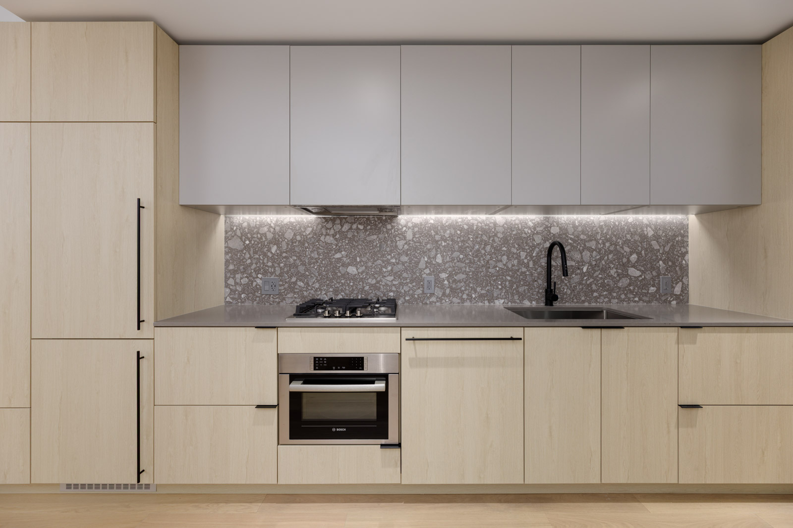 Modern kitchen with light wood cabinets, gray countertops, built-in oven, gas stove, gray upper cabinets, and a stone-patterned backsplash. A sink with a black faucet is on the right.
