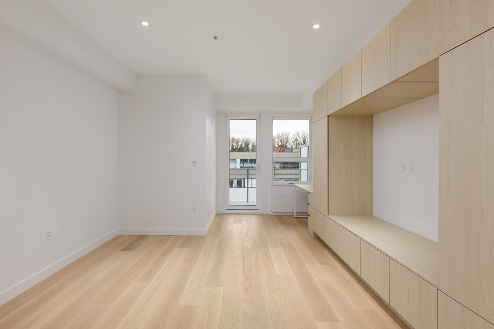 Empty modern room with light wood flooring, built-in light wood cabinetry along one wall, white walls, ceiling lights, and a glass door leading to a balcony with an outdoor view.