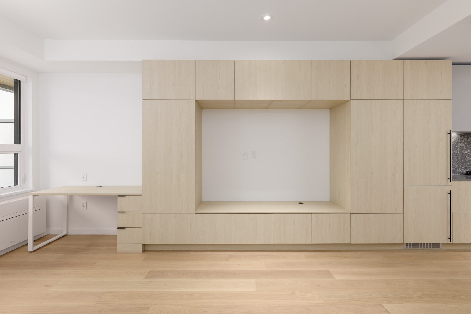 Minimalist room with light wood built-in cabinetry, an integrated desk on the left, and an empty recessed space in the center, set against white walls and light hardwood floors.