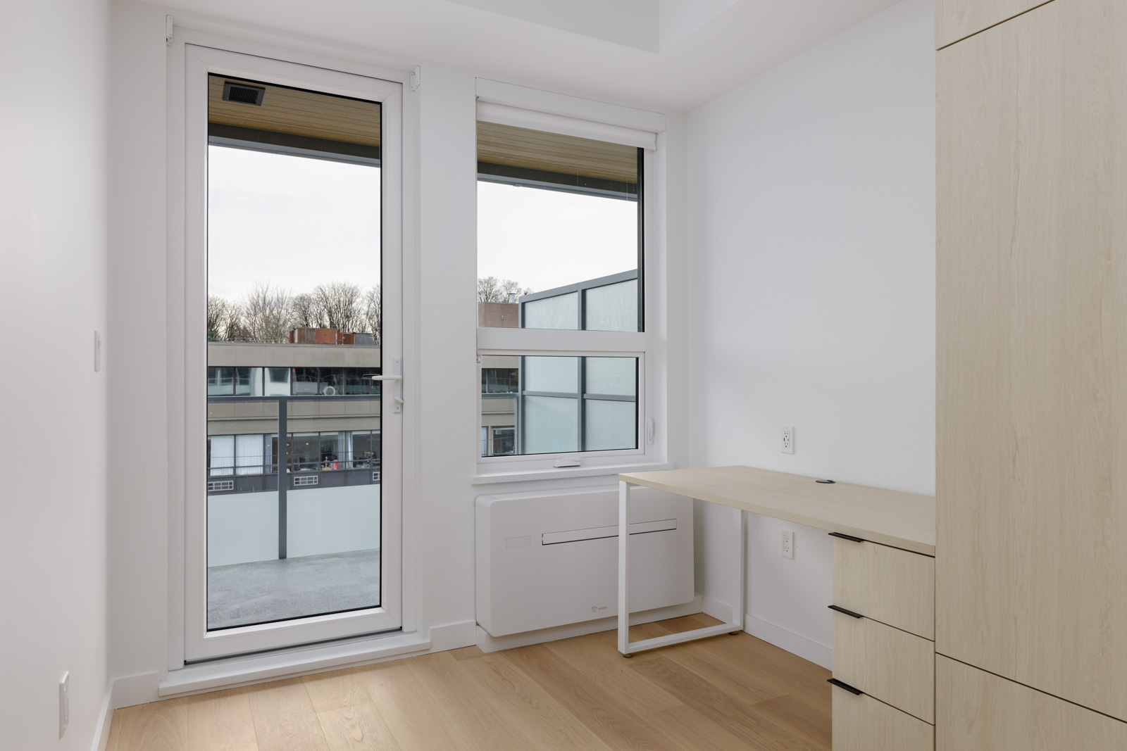 A small, modern room with light wood flooring, a built-in wardrobe, a minimalist desk, and large windows overlooking nearby buildings.
