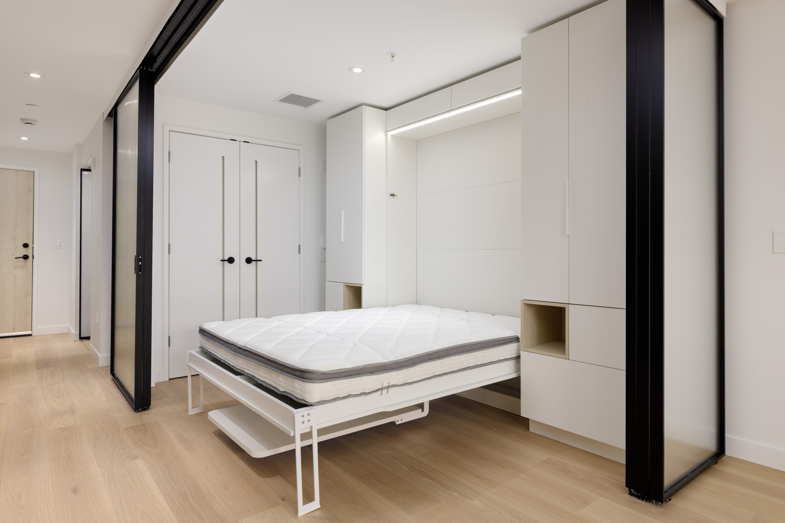 A modern room with light wood flooring and a pull-down Murphy bed extended from a white built-in wall unit, flanked by sliding glass doors.